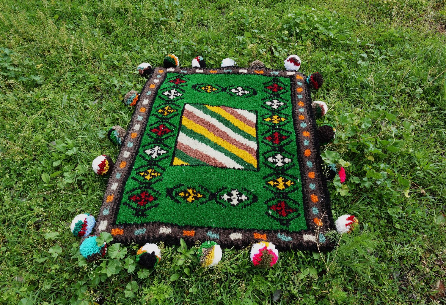 Hand-woven green Shabrak with geometric pattern and pompoms on grass