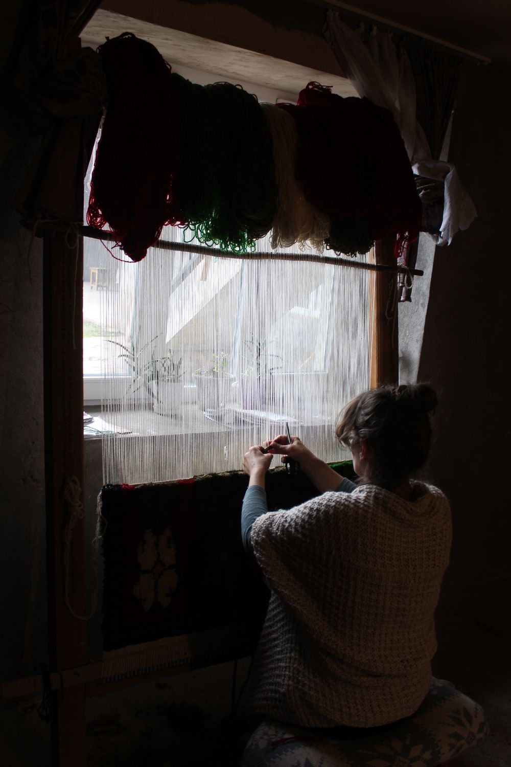 The weaver at her wooden loom by a window with dyed wool hanging above